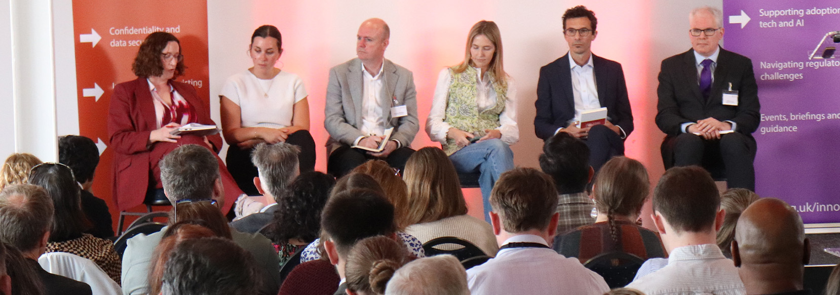 A panel of six people is seated on a stage at a conference. They are facing an audience. The backdrop features text related to navigating regulatory challenges and technology. Audience members are visible in the foreground.