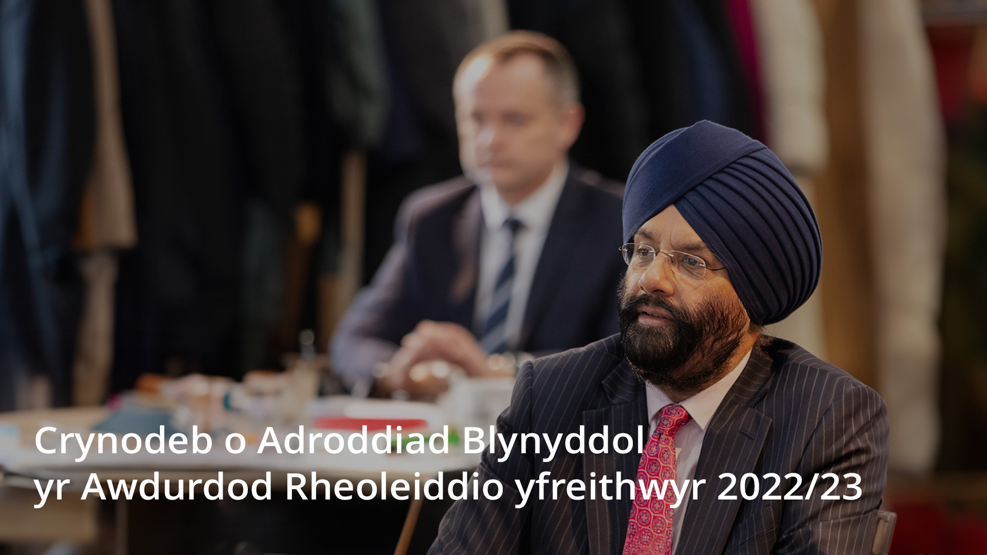 Person wearing a turban and glasses sitting thoughtfully at a conference table, with another individual in the background. A button reading 'Read the summary report' is displayed prominently in the foreground.